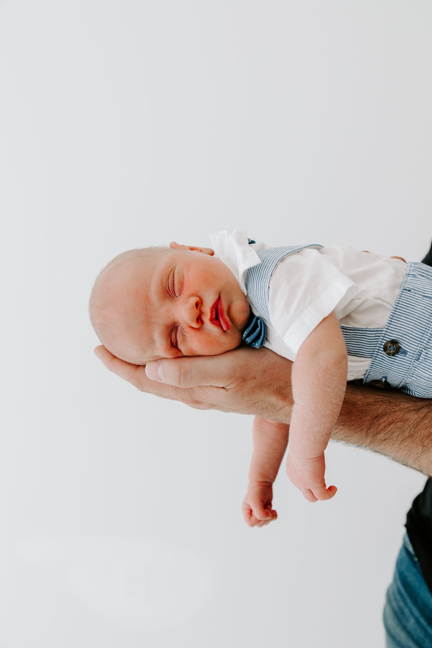 Sleeping baby in blue and white outfit cradled gently in someone's arms.