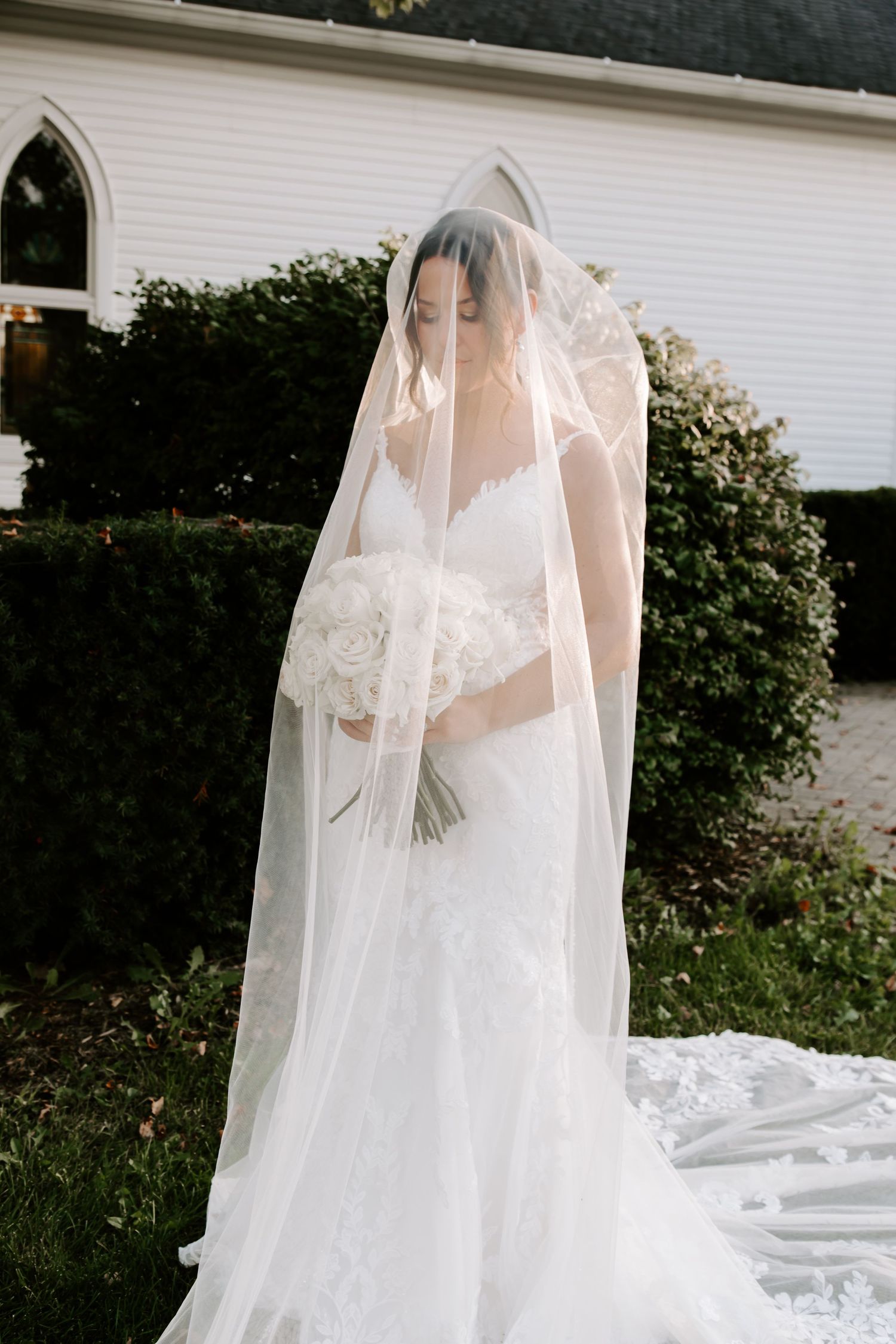 Ethereal bride in white lace dress with flowing cathedral veil against white church backdrop.
