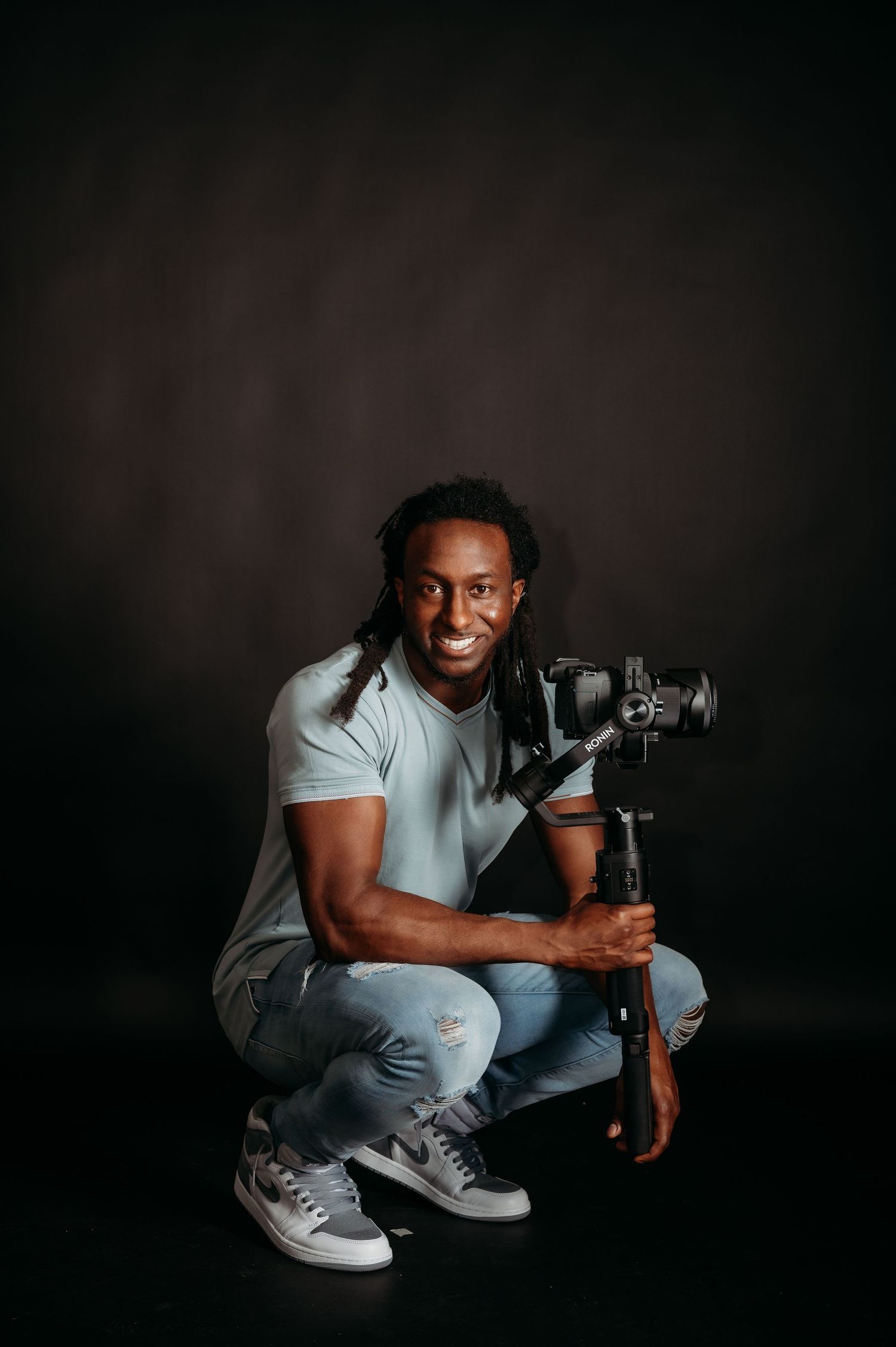 A photographer in a light colored shirt and jeans crouches with professional camera equipment against a dark backdrop.