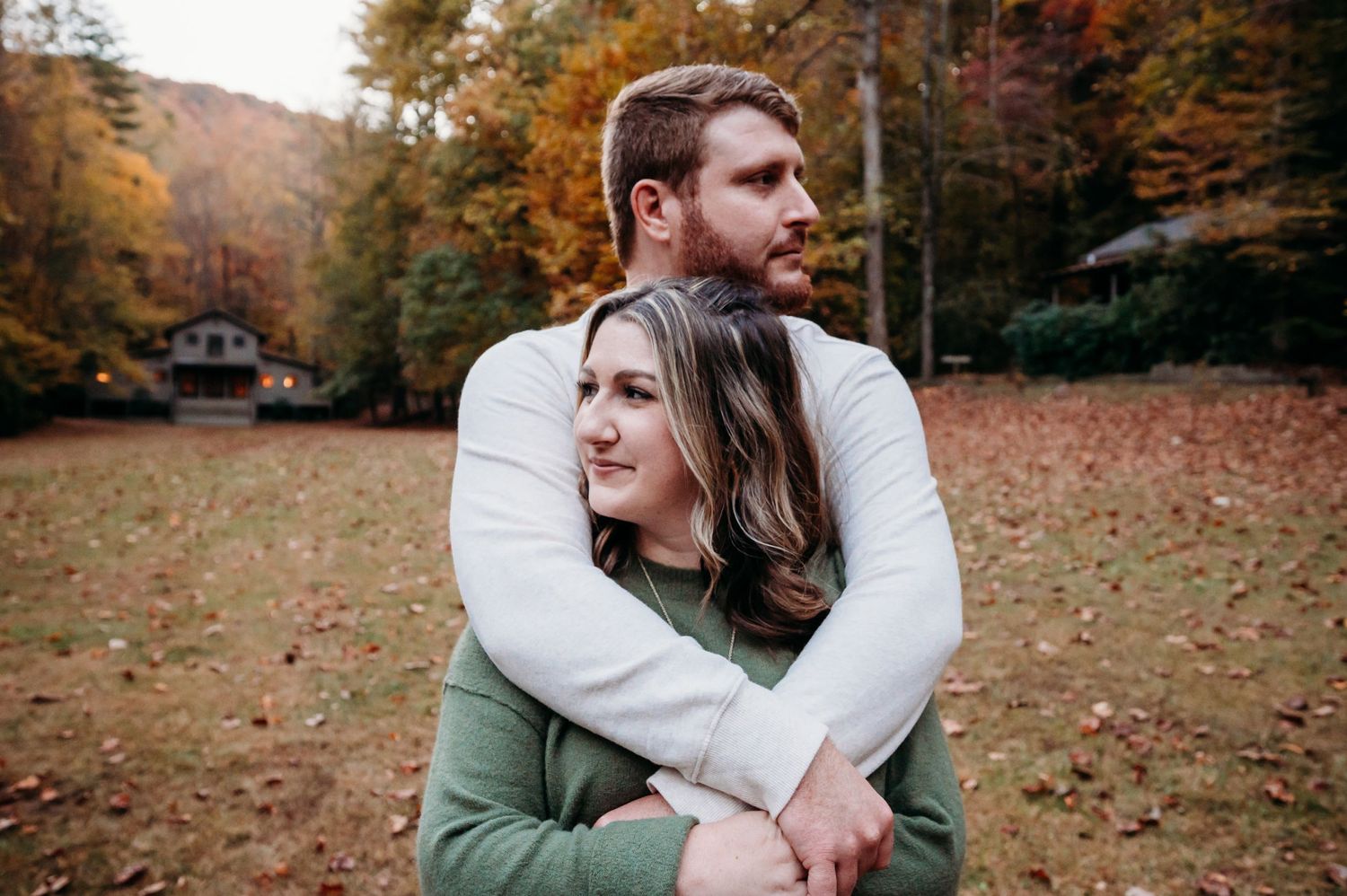 A couple embraces in a cozy embrace while sitting in a fall forest clearing with autumn foliage in the background.