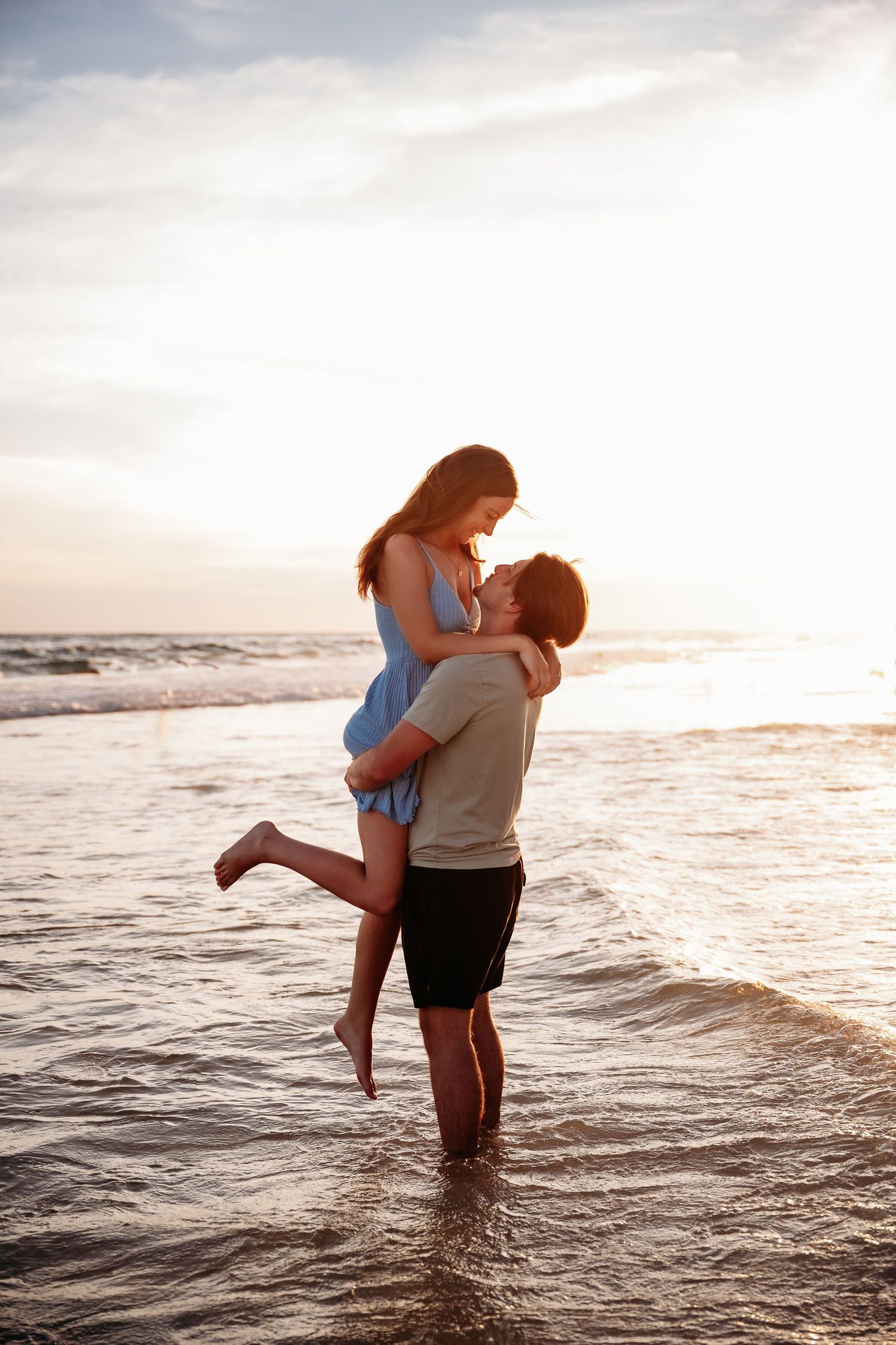 A romantic couple embraces while standing in shallow ocean waves at sunset on the beach.