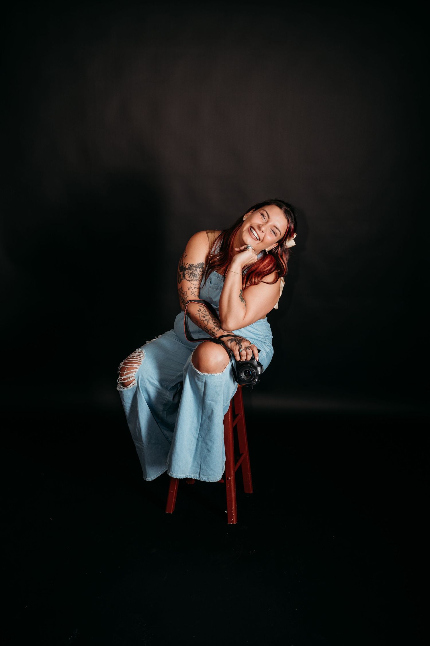 Artistic portrait of someone wearing ripped jeans and a tank top posing on a red stool against a dark background.