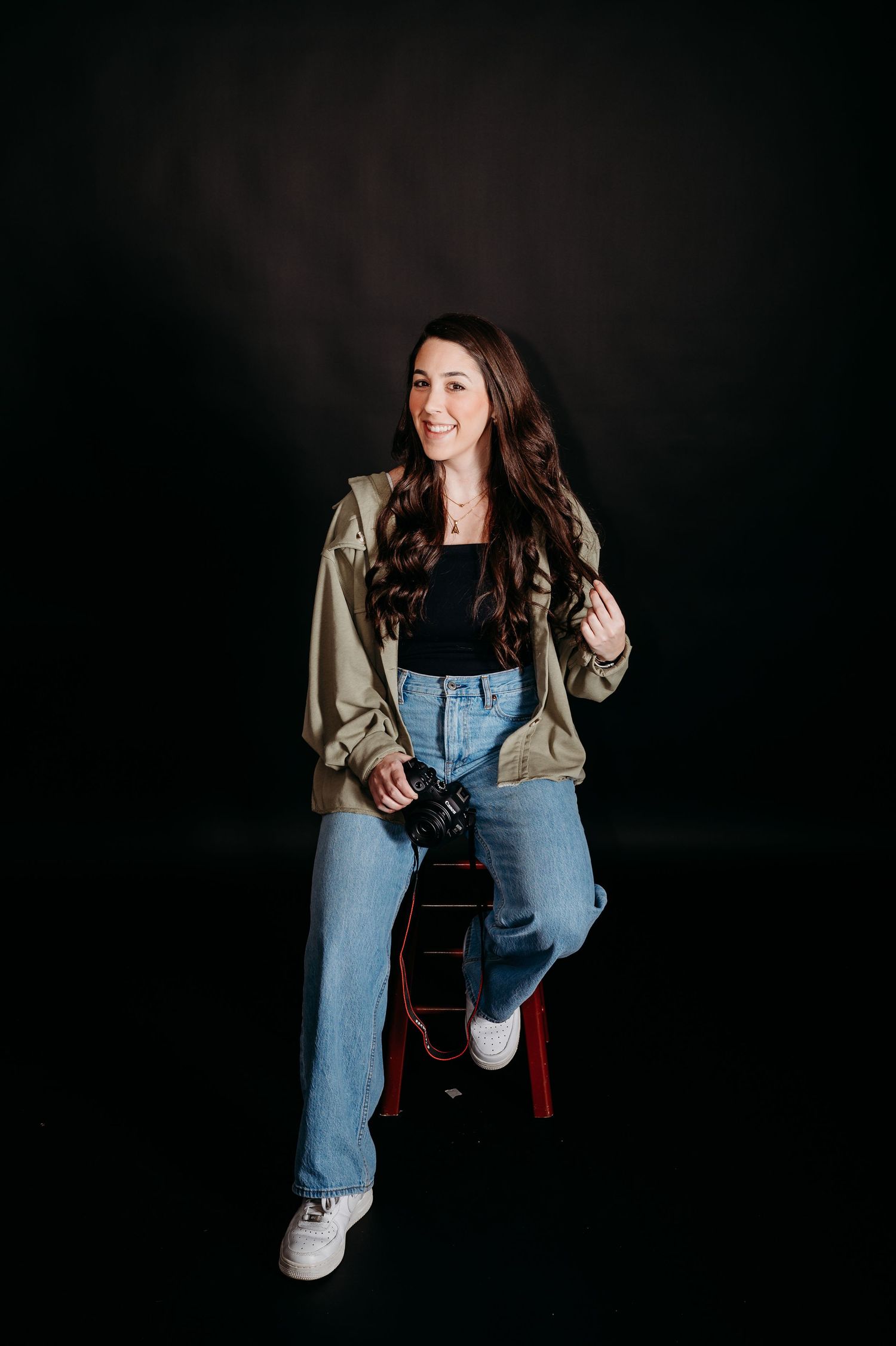 Portrait against dark background showing casual 90s style outfit with baggy jeans, black top and tan bomber jacket.