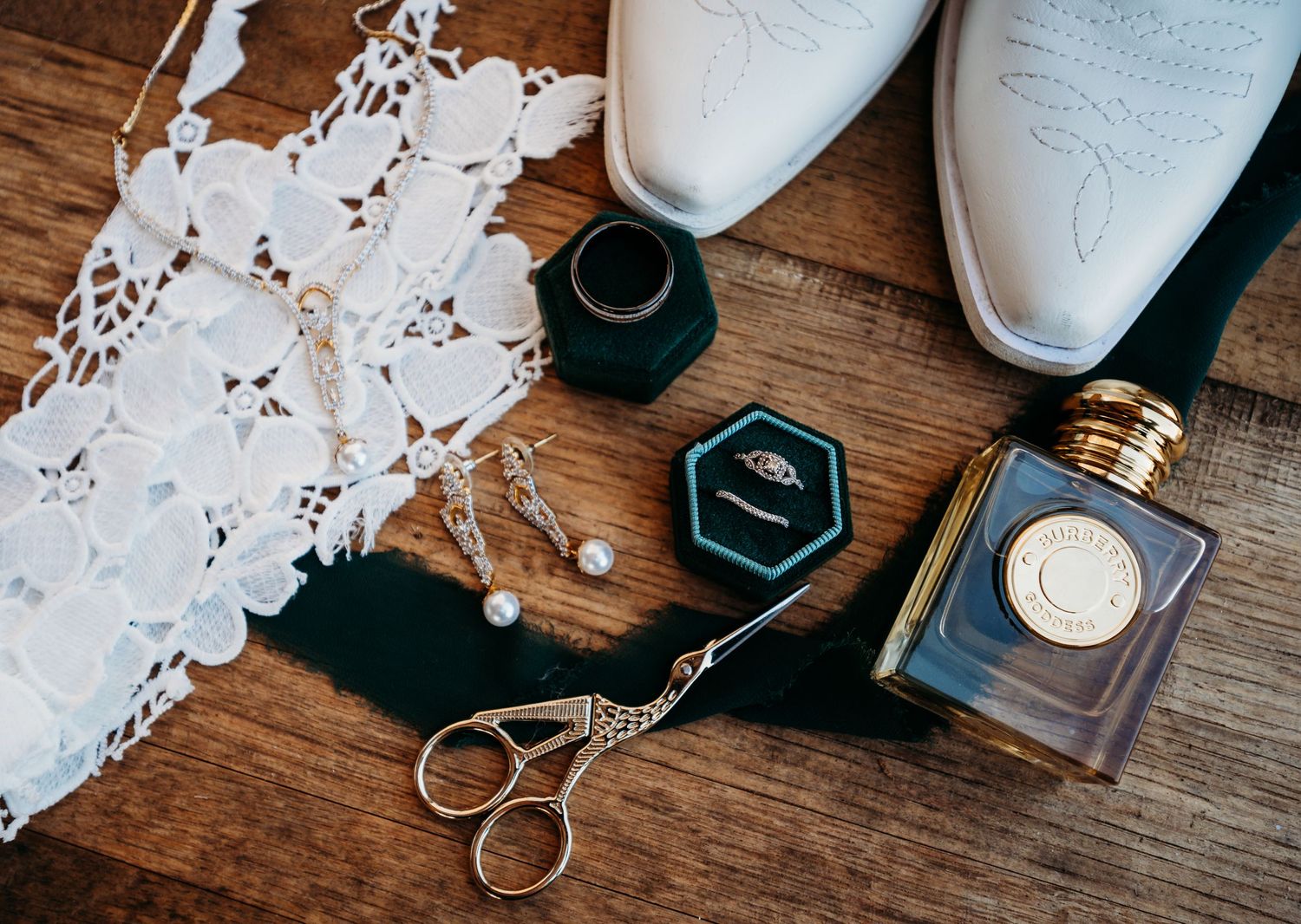 Wedding details flat lay featuring white boots, vintage scissors, lace trim and perfume bottles on wooden surface.