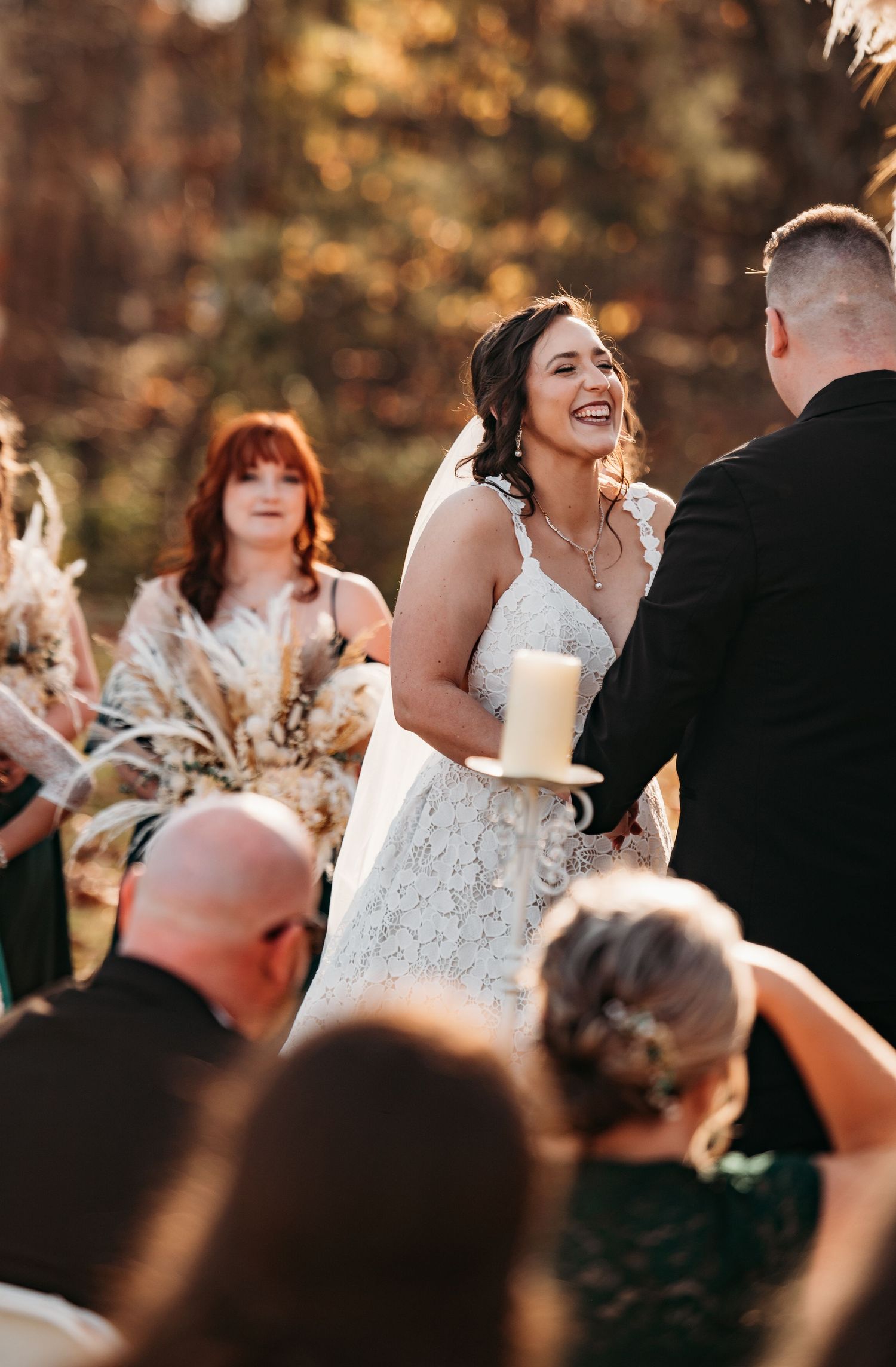 A joyful outdoor wedding ceremony shows a bride in a white lace dress sharing a candid laughing moment during the service.