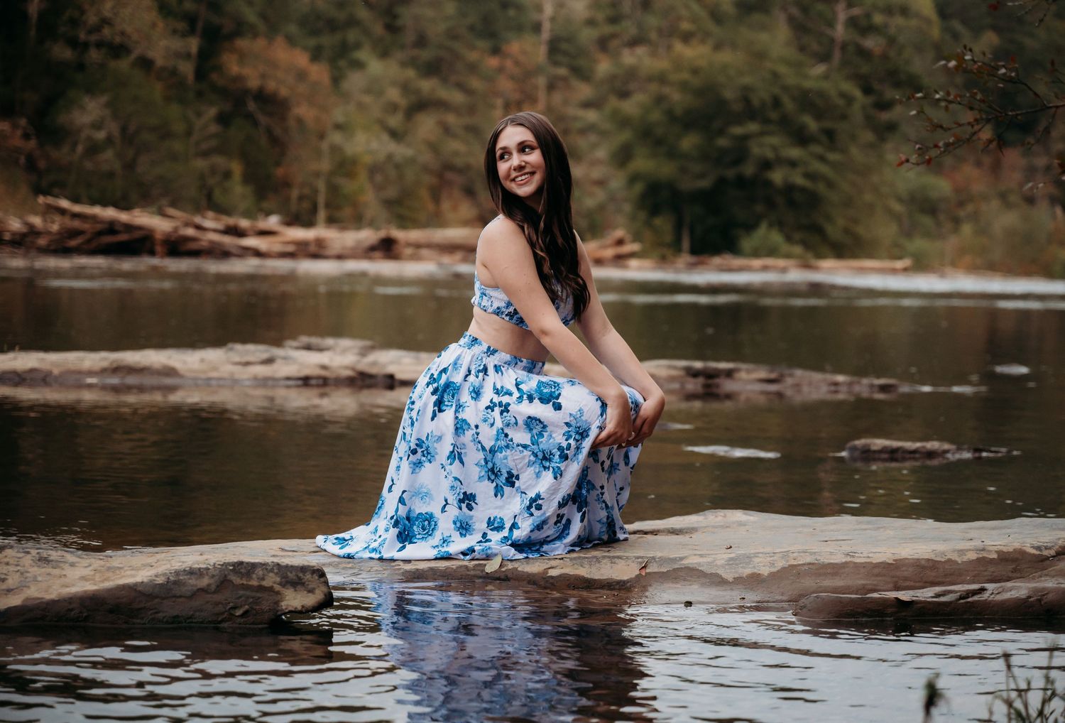 A person in a floral blue dress poses on rocks by a serene river during autumn.