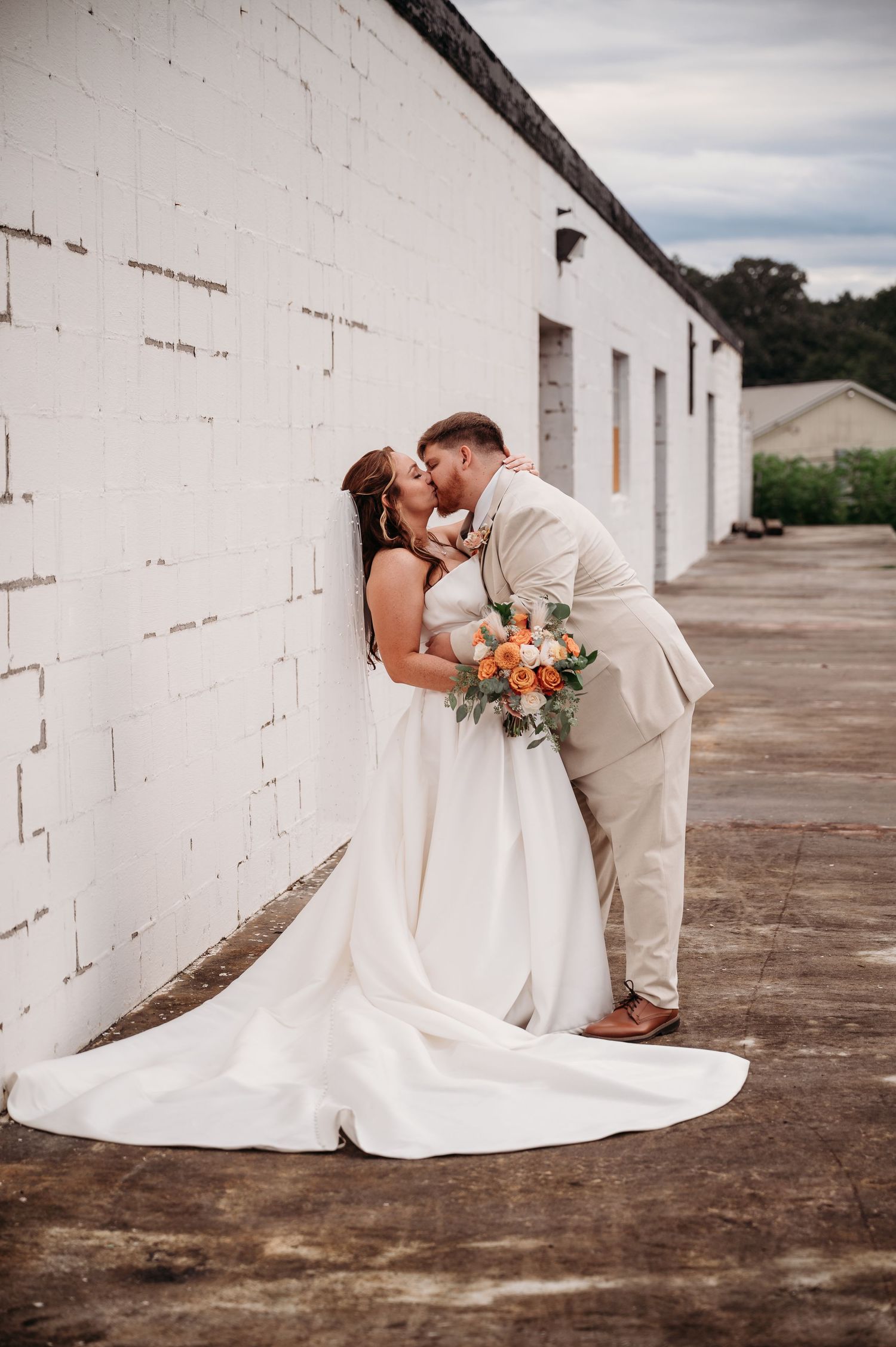 A couple shares a romantic kiss on their wedding day against a white brick wall, with the bride holding a peach floral bouquet.
West Ga, FL