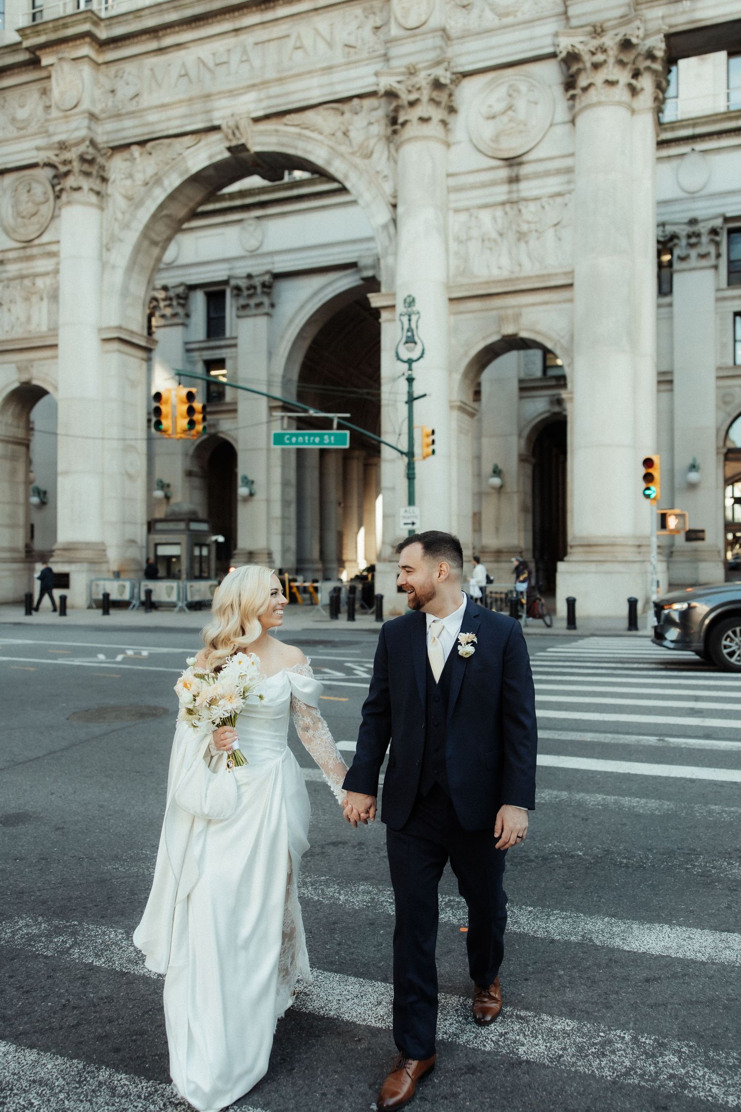 A couple in wedding attire walks together on a city street with grand architectural buildings in the background.