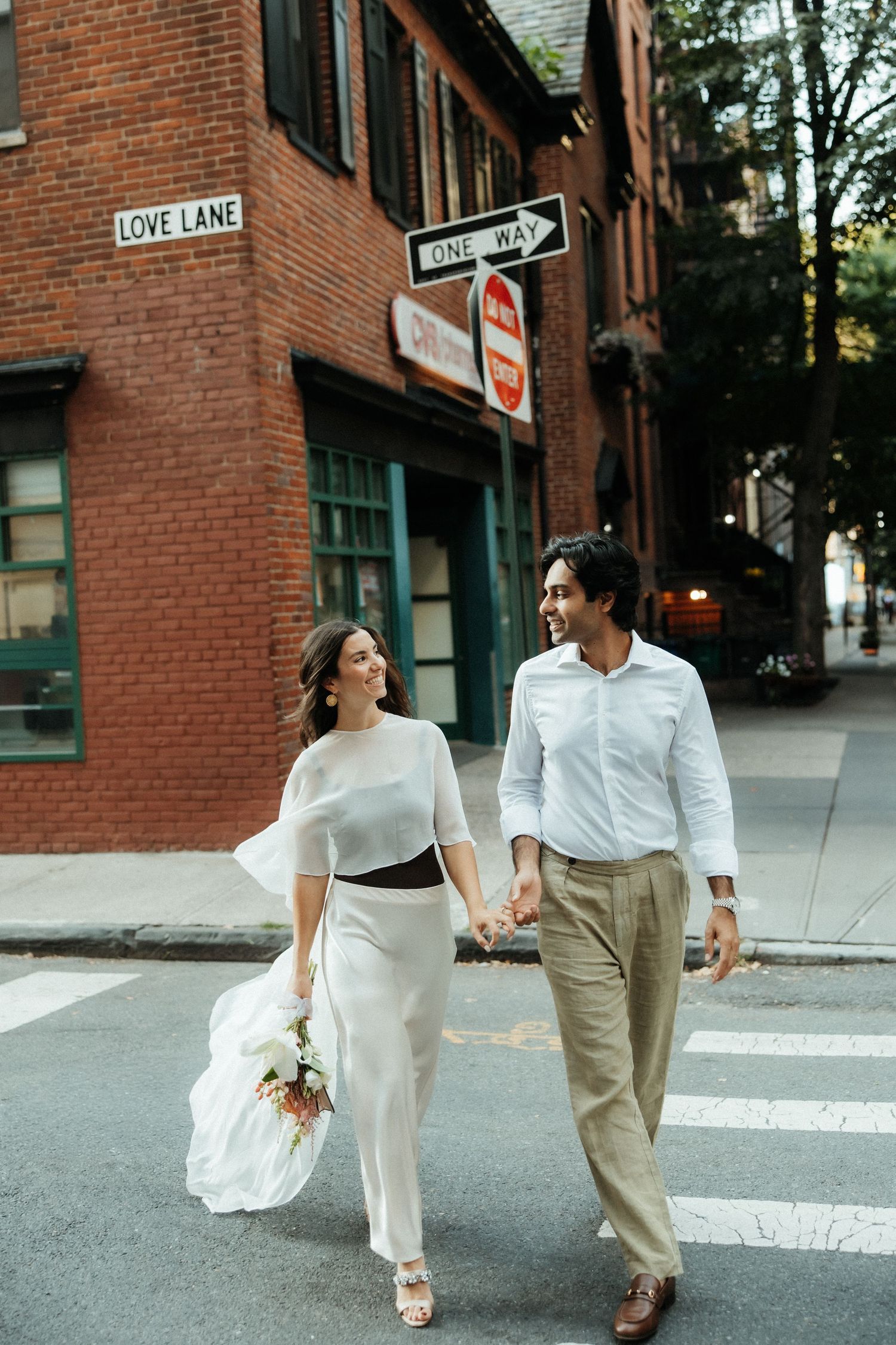 A couple walks hand in hand on Love Lane in a charming brick neighborhood, with the bride holding a bouquet.