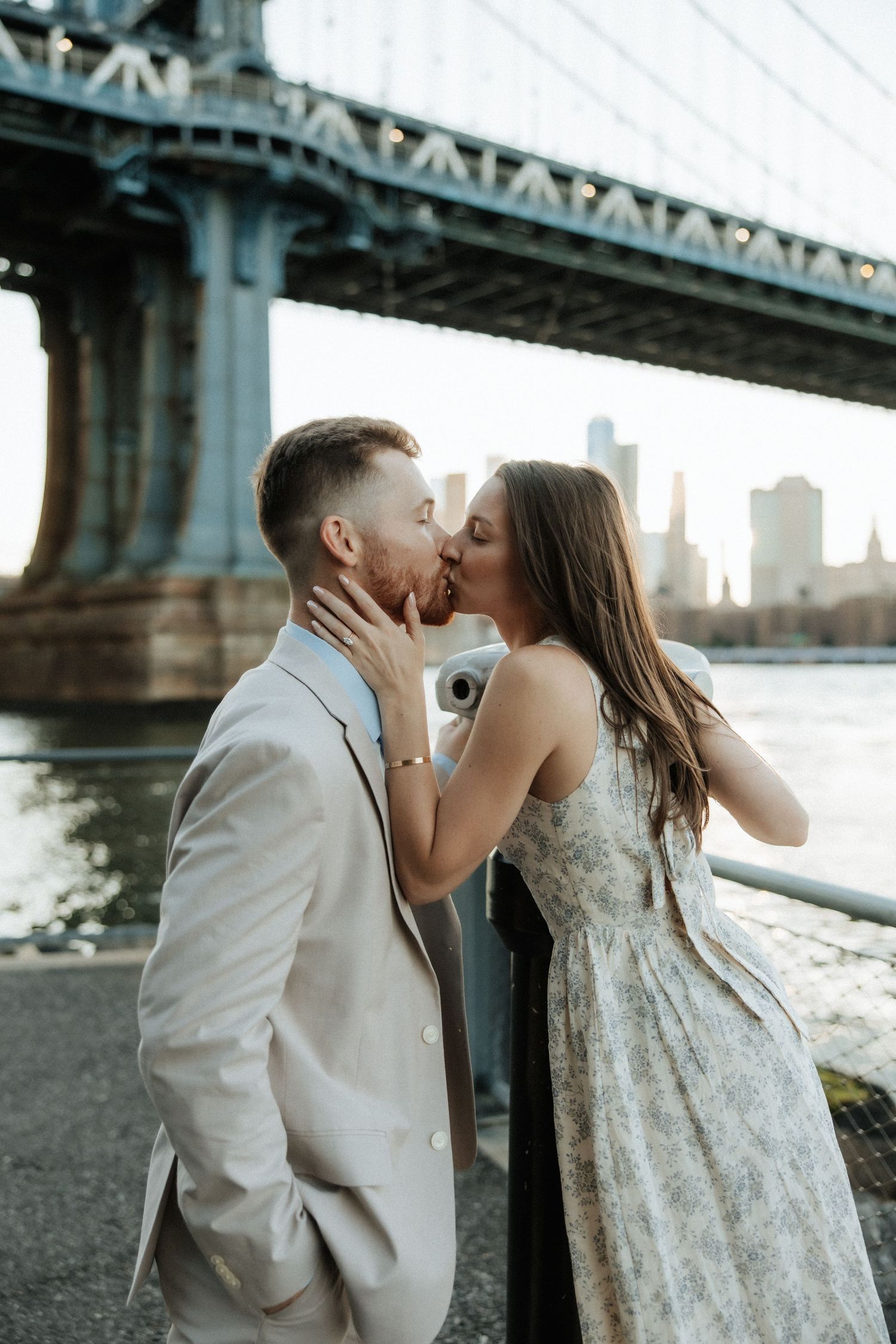 A romantic couple shares a kiss under the Manhattan Bridge in New York City during sunset.