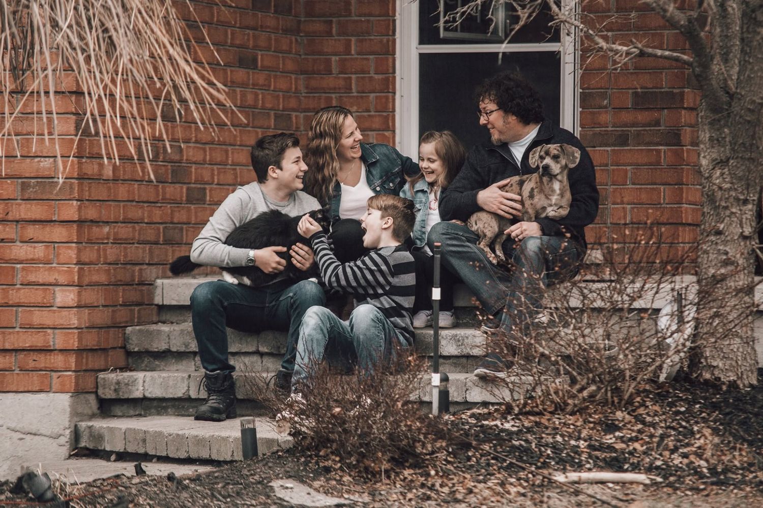 Family with two dogs sitting on brick home front steps, laughing together outdoors in casual winter clothing.