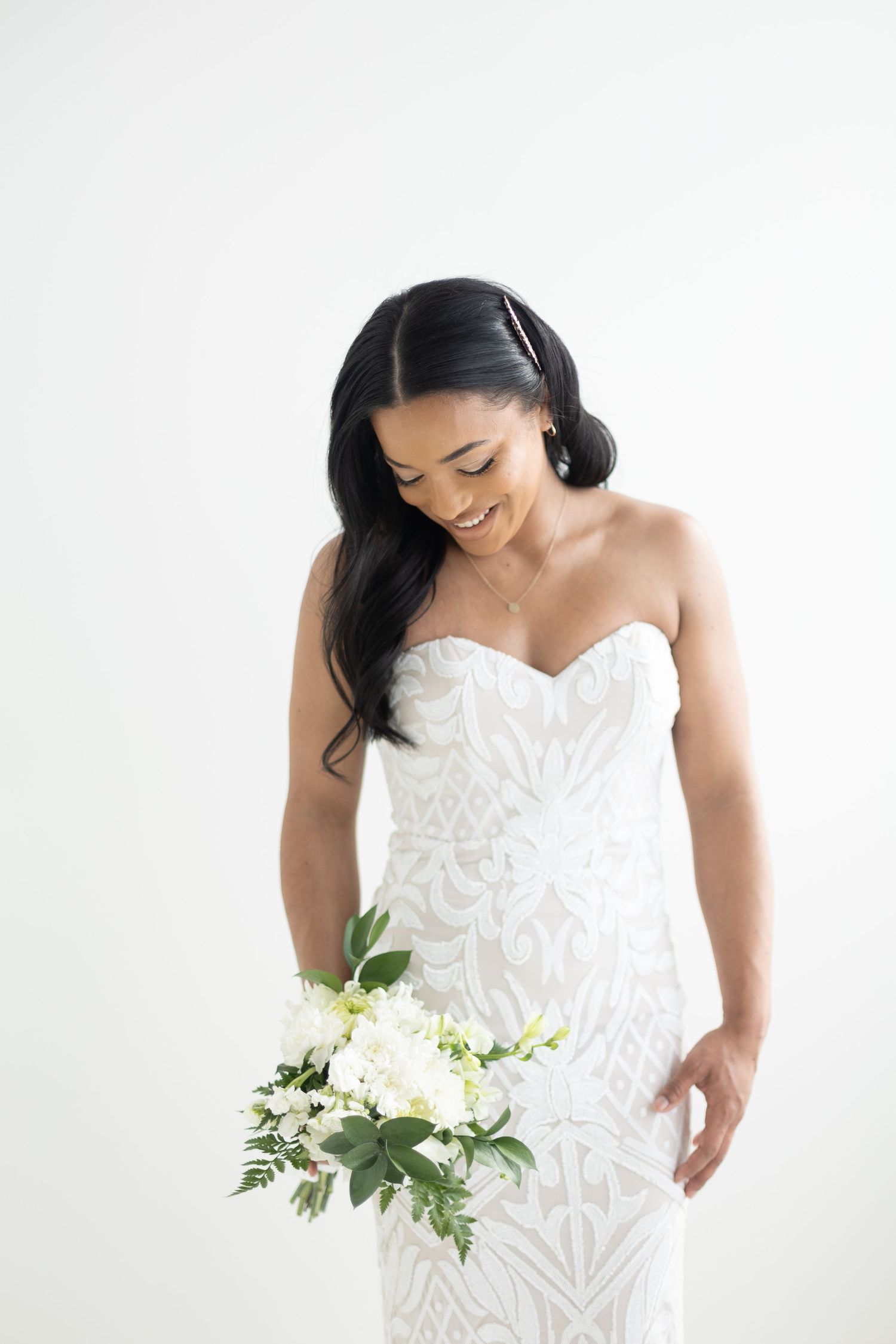 Elegant bride in strapless lace gown smiling down at white floral bouquet.