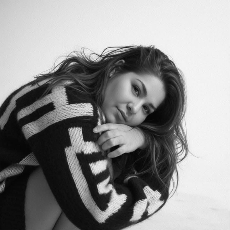 Black and white photo of a young woman with wavy hair wearing a bold patterned knit sweater, posing gracefully.