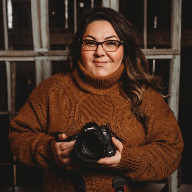 Woman in brown cable-knit sweater and glasses holds Canon DSLR camera, posing in front of rustic window backdrop.