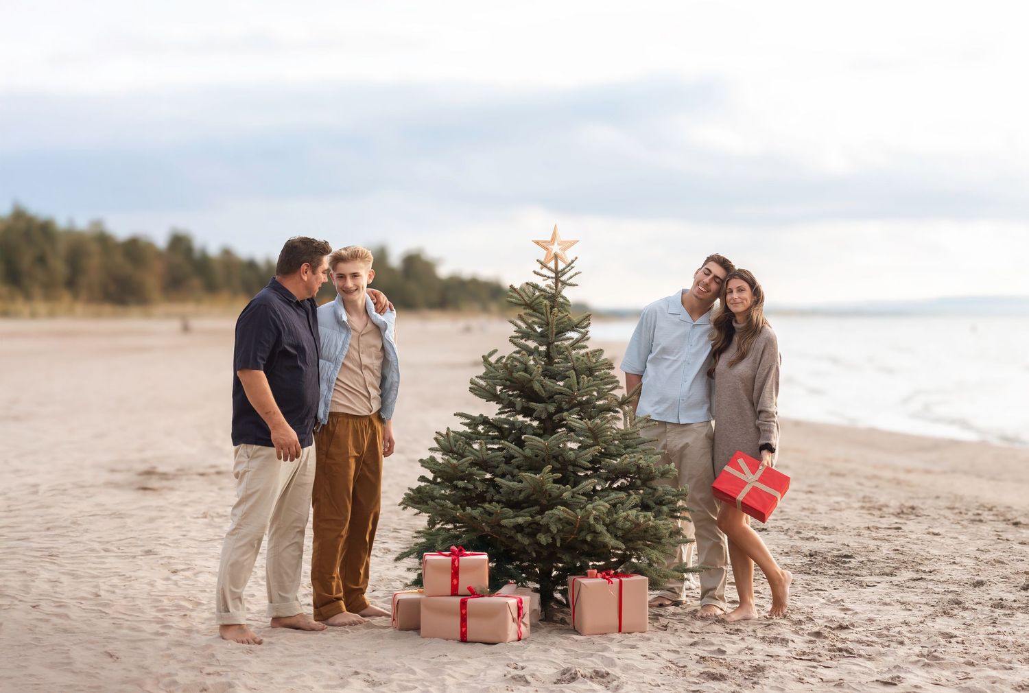 Four people celebrate Christmas on a beach with a decorated tree and wrapped gifts in the sand.