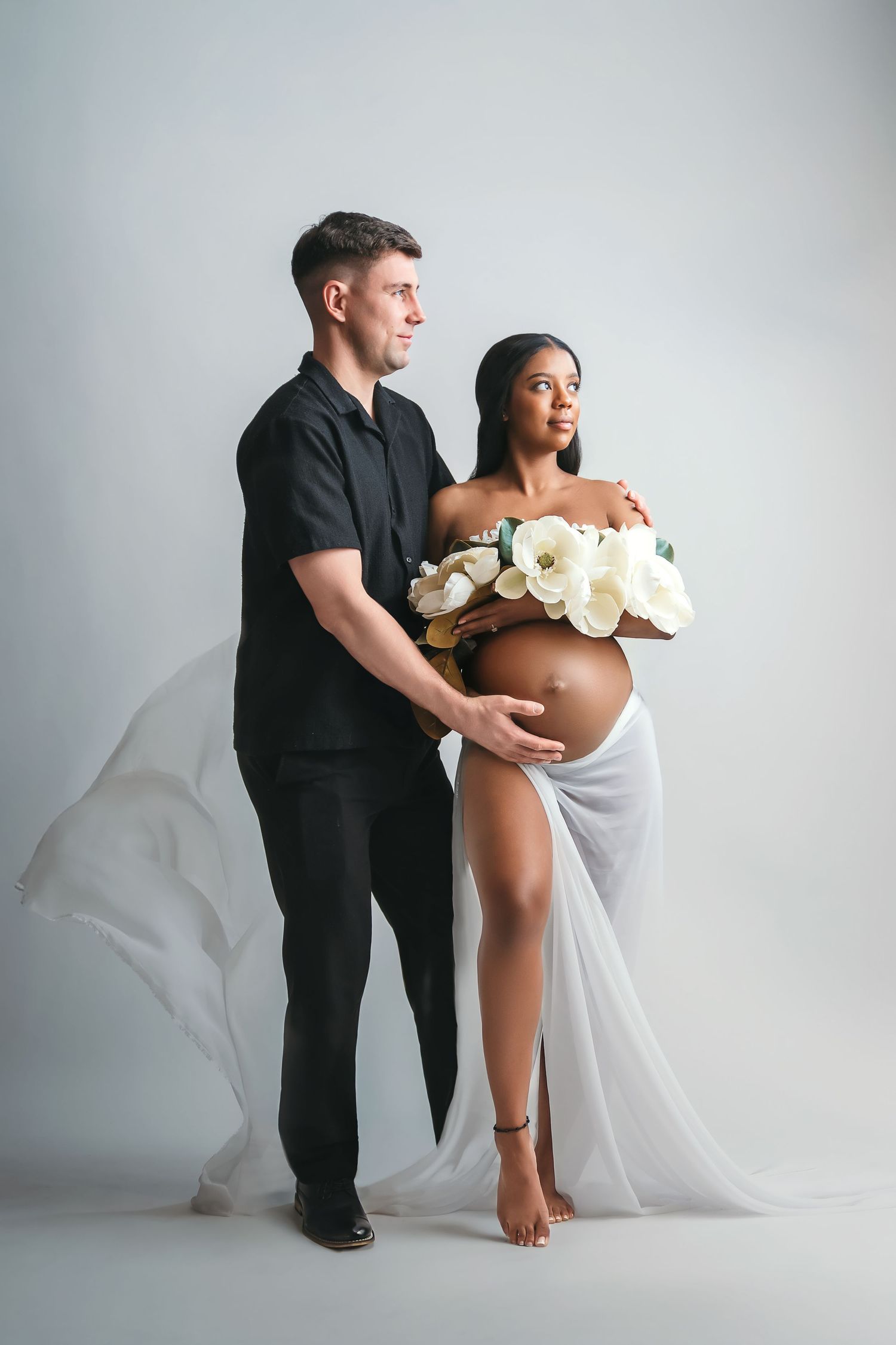 Interracial couple maternity photo, man in black shirt embracing pregnant woman with white flowers and flowing fabric.