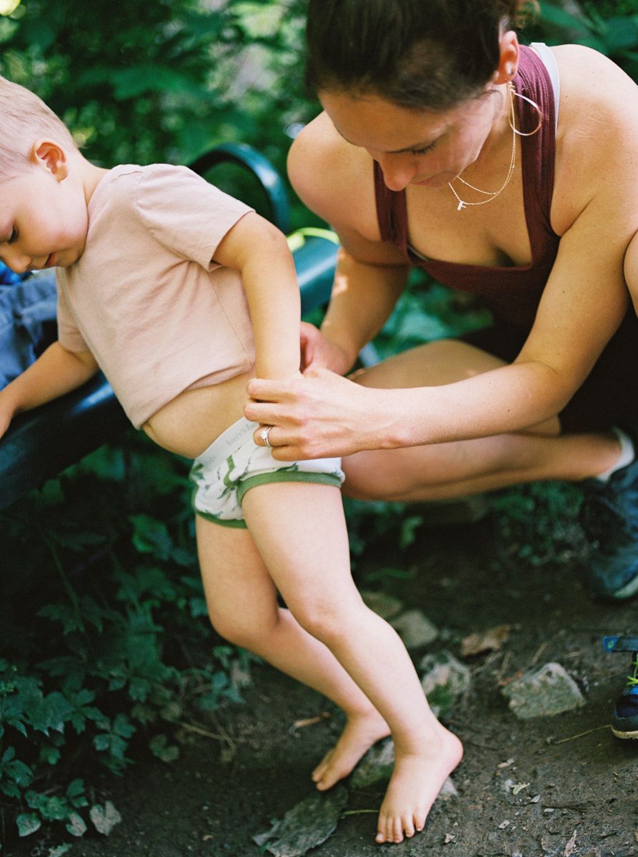 A woman crouches outdoors helping a young toddler with their diaper amid lush green foliage.