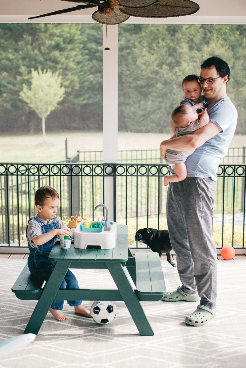 Father holding twin babies while toddler plays at outdoor picnic table on porch.