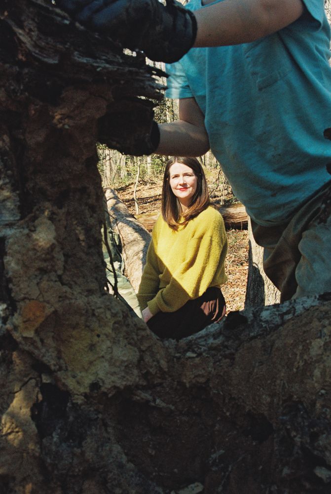 Creative framing through a hollow tree trunk shows a person in a bright yellow sweater standing in a wooded area.