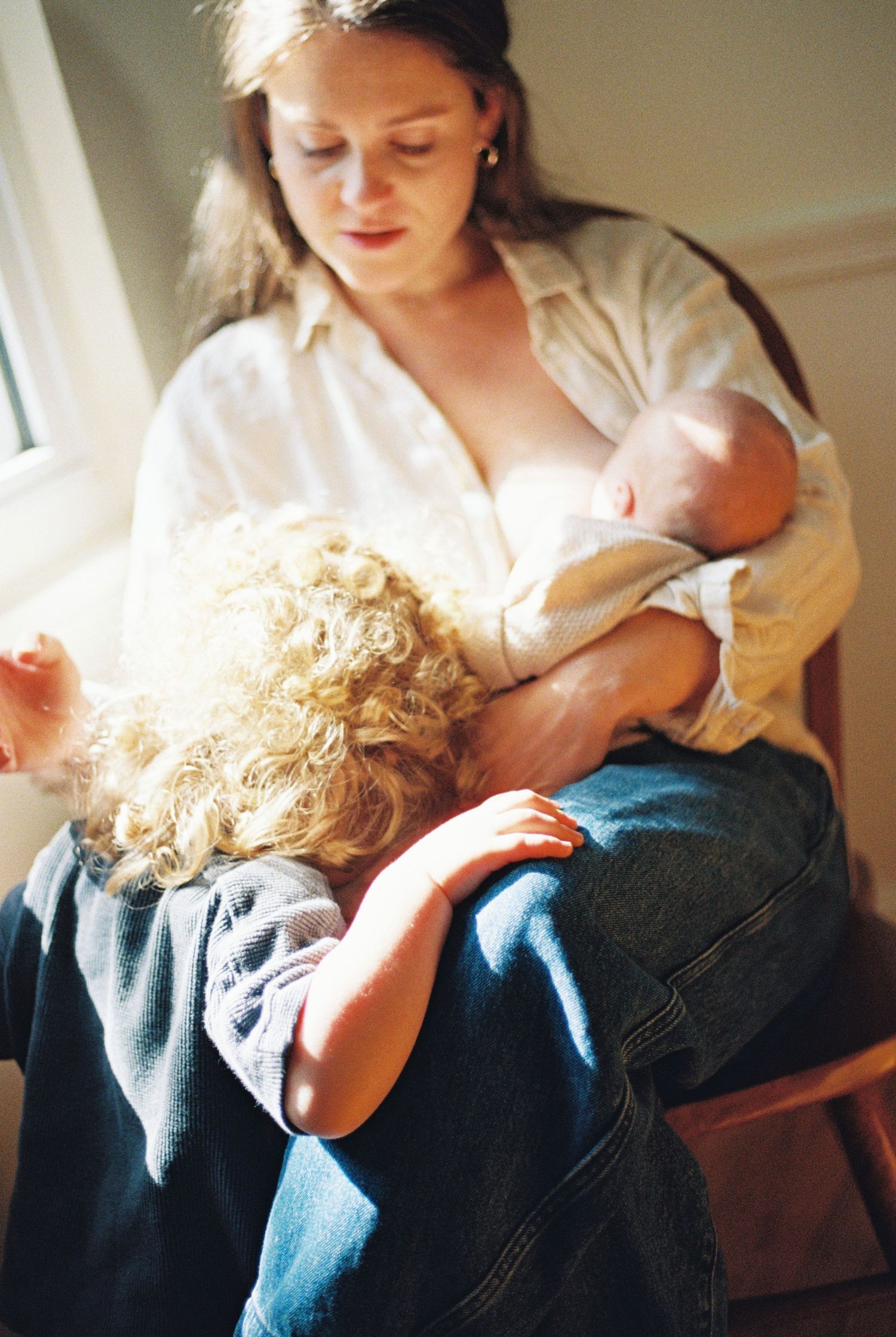 Mother breastfeeding newborn baby while toddler with curly blonde hair rests on her lap near sunny window.