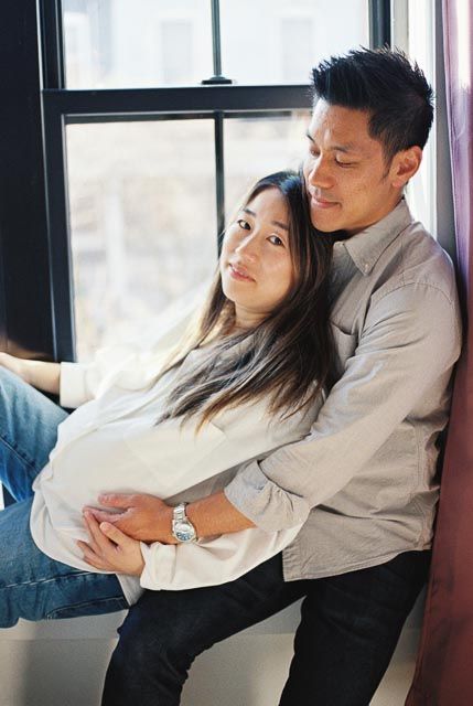 Pregnant Asian woman embraced by man near window, maternity couple portrait.