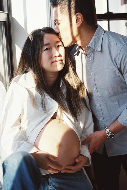 Pregnant Asian woman in white shirt holds her belly while man in blue shirt kisses her forehead near a window.