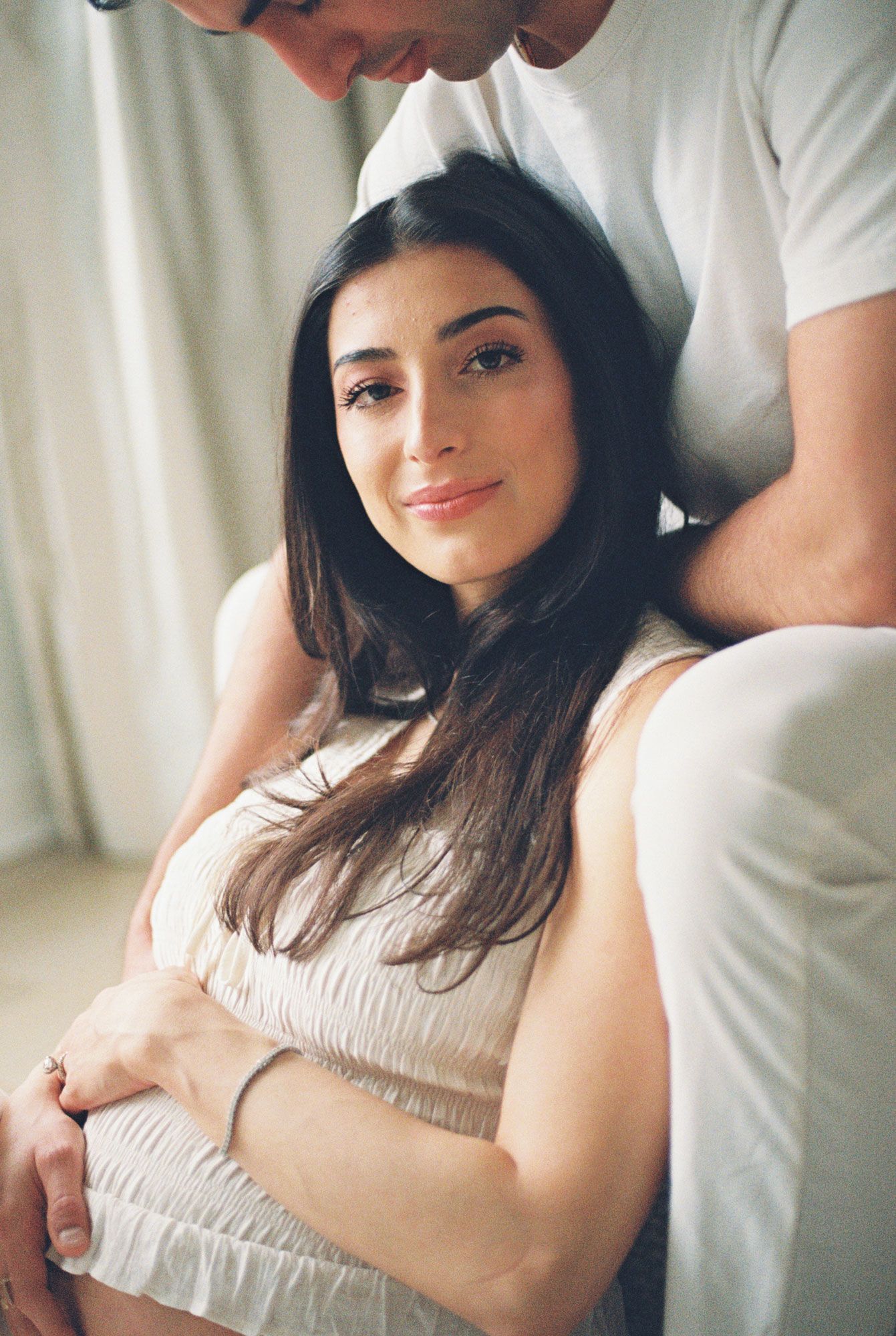 Smiling pregnant woman with long dark hair rests her hands on her belly while her partner embraces her from behind in soft natural light.