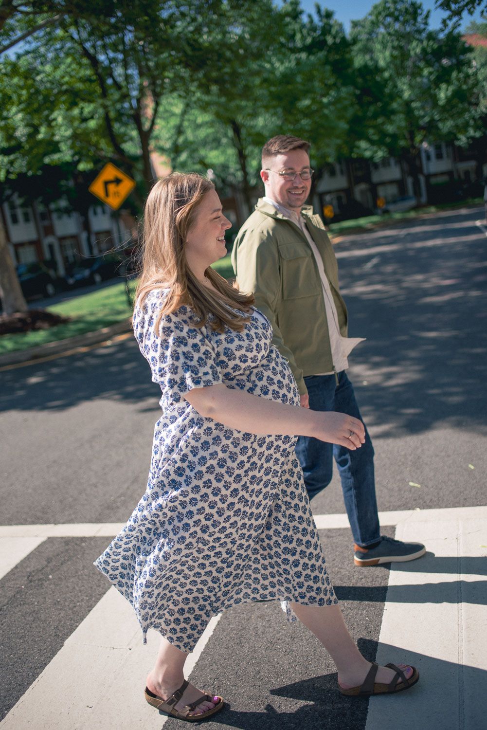 Pregnant woman in floral dress and man in green shirt crossing a sunny street crosswalk, smiling and laughing together.