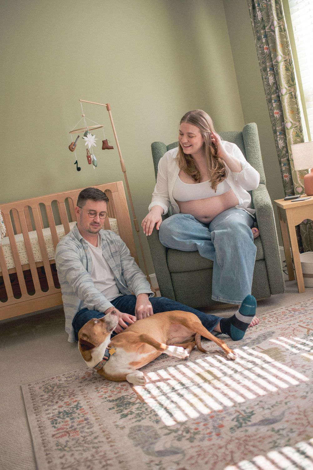 Expectant couple relaxing in nursery with dog, pregnant woman in glider chair beside wooden crib.