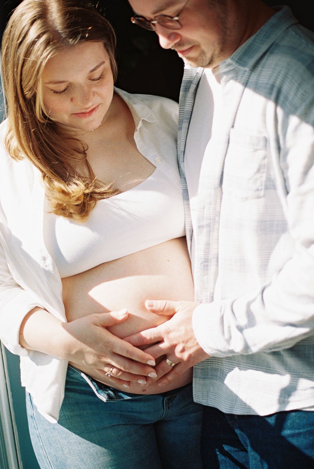 Couple tenderly holding pregnant woman's baby bump together near sunny window.