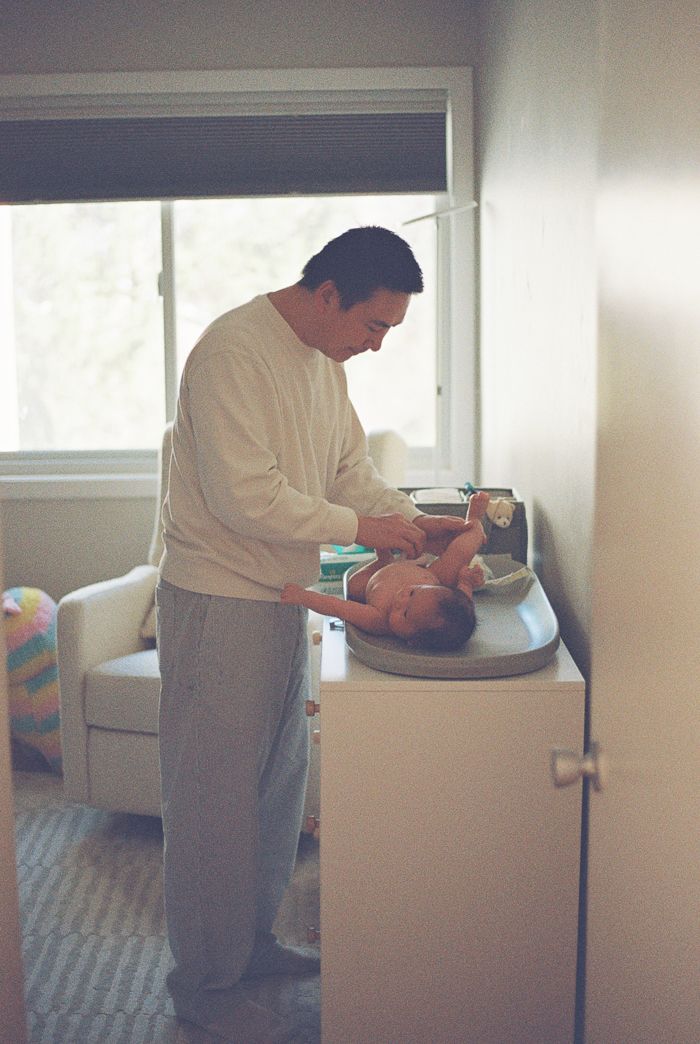 A father in a white sweater changes his newborn baby's diaper on a changing table by a bright window.