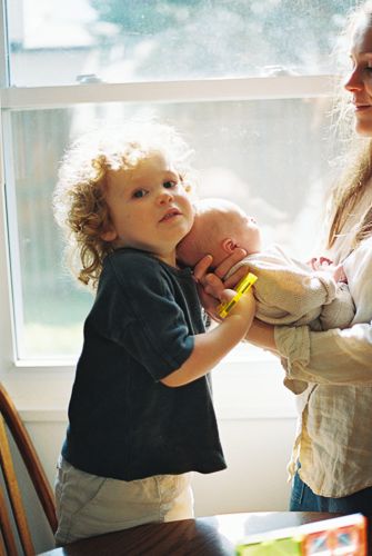 Curly-haired toddler holding a newborn baby near a bright window with an adult nearby.