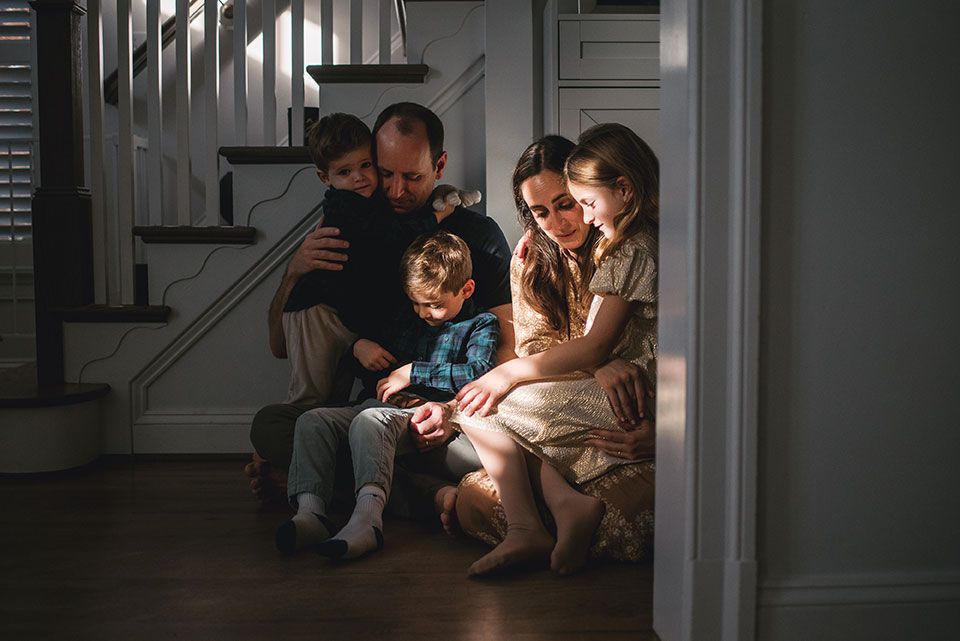 Family sits together on stairs in a dimly lit home interior looking at glowing digital device screen.