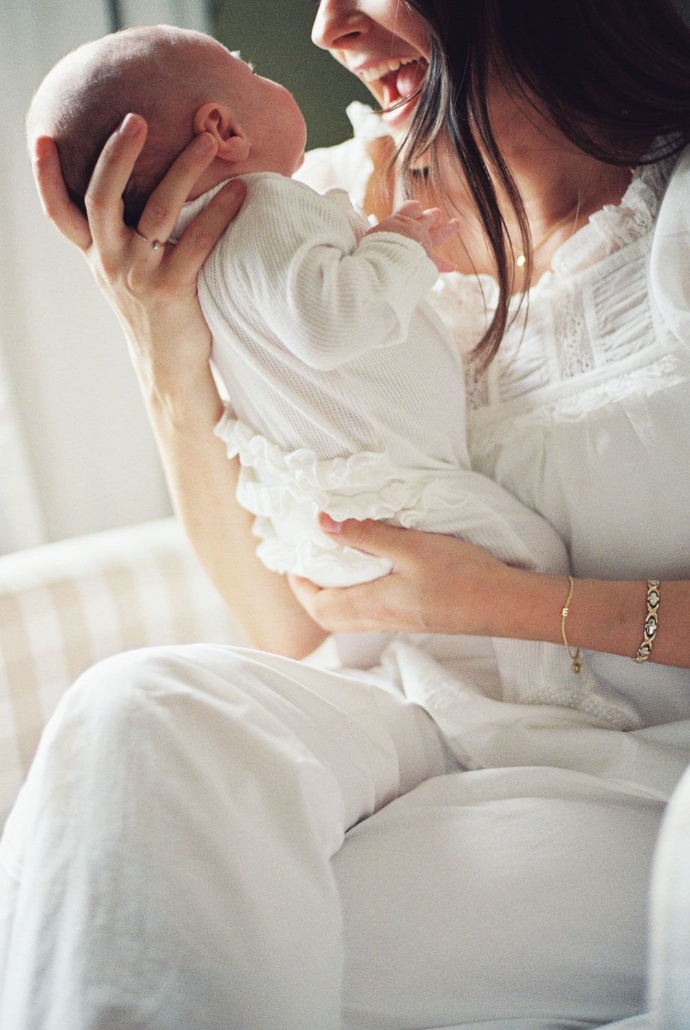 Mother in white dress joyfully lifts her newborn baby, both dressed in white, in a bright nursery setting.