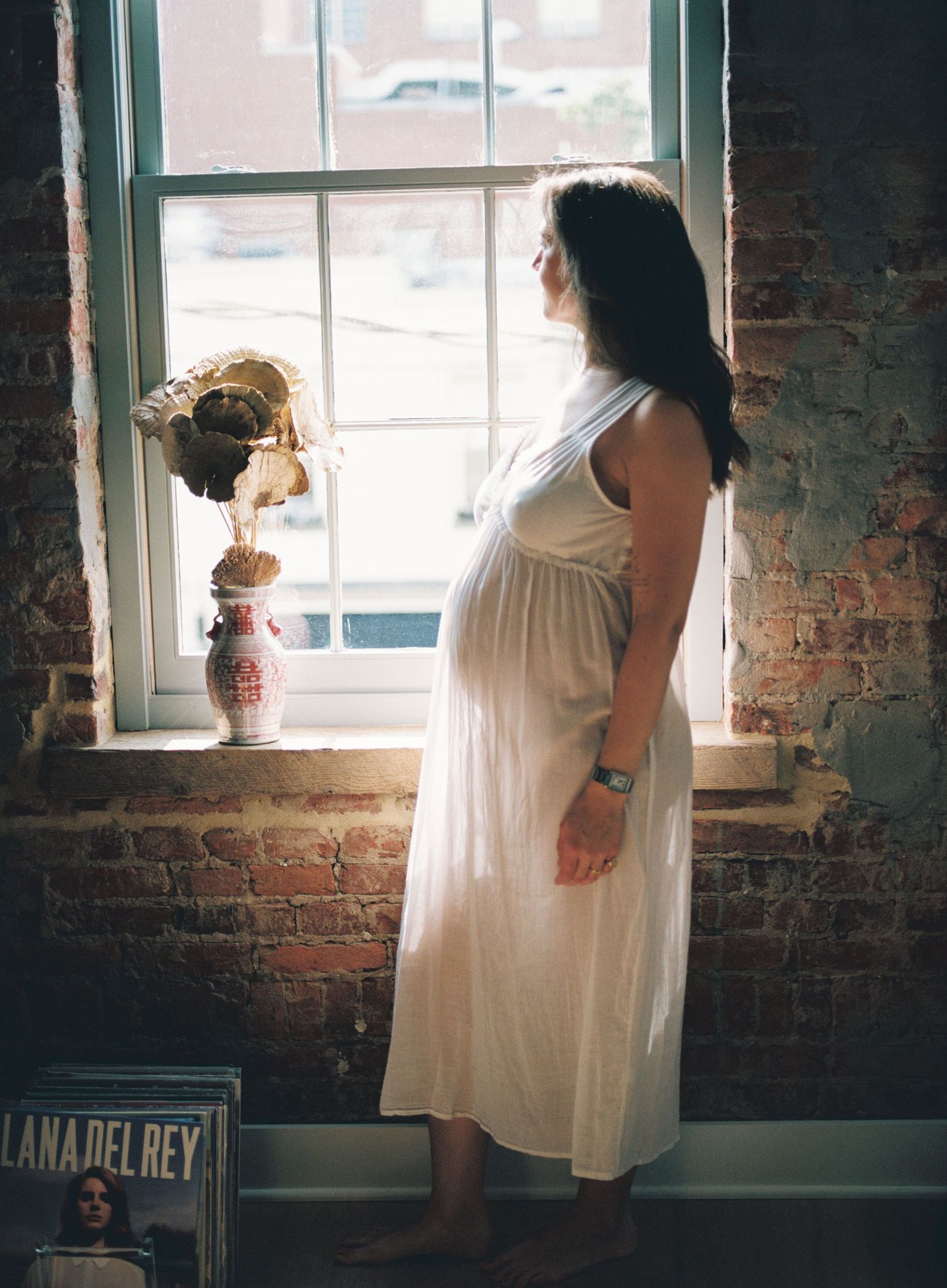 Pregnant woman in white dress gazing out a window beside a brick wall and floral vase.