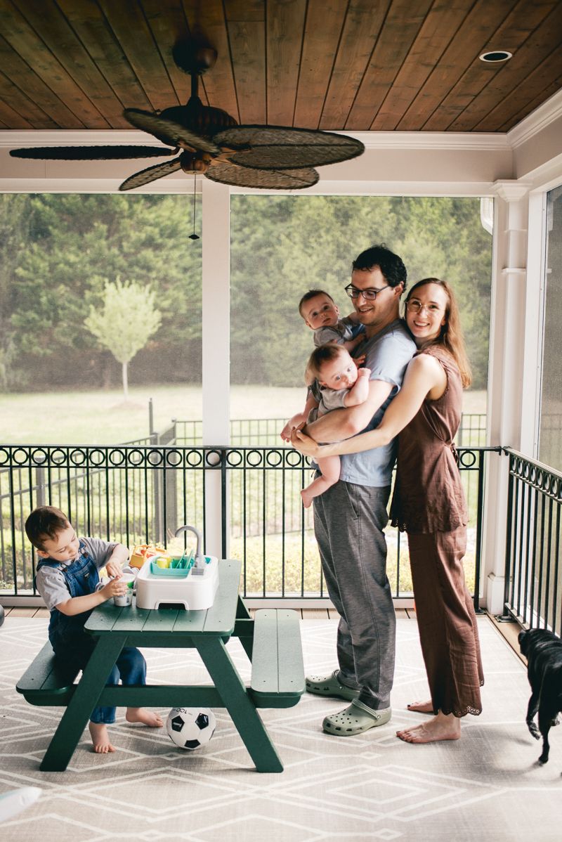 Family of five posing on a covered porch with ceiling fan, green picnic table, and lush backyard views.