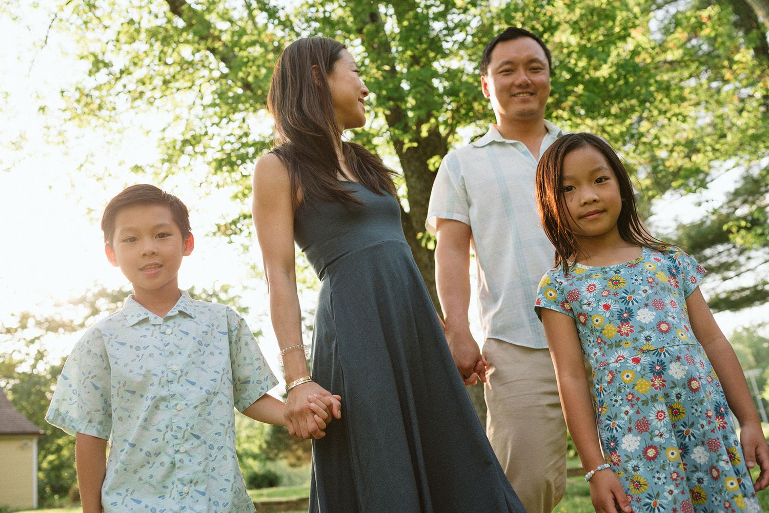 A family group holds hands while standing together outdoors under green trees on a sunny day.
