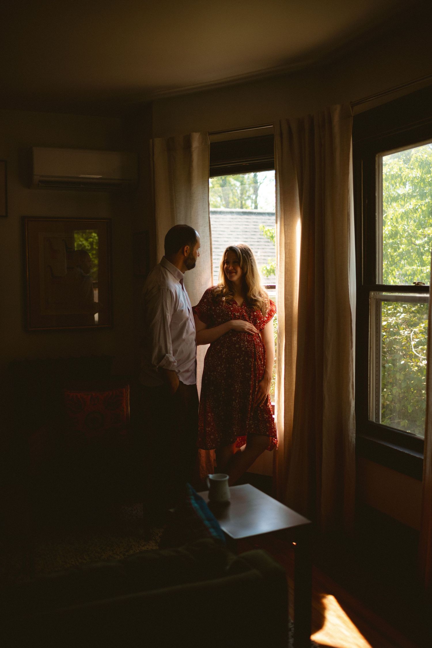 A couple stands together near a window, silhouetted by warm natural light streaming through sheer curtains.