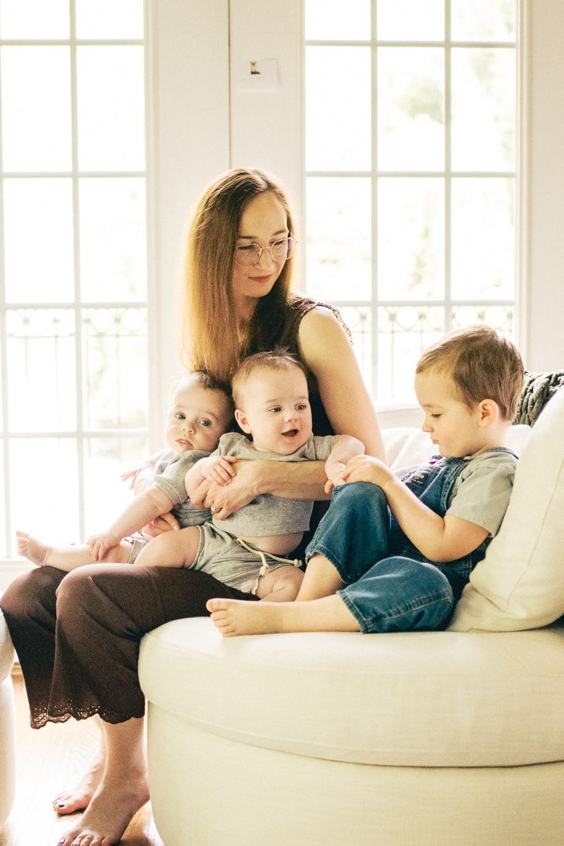 A young mother sits on a white sofa holding two babies while a toddler boy sits beside her near a bright window.