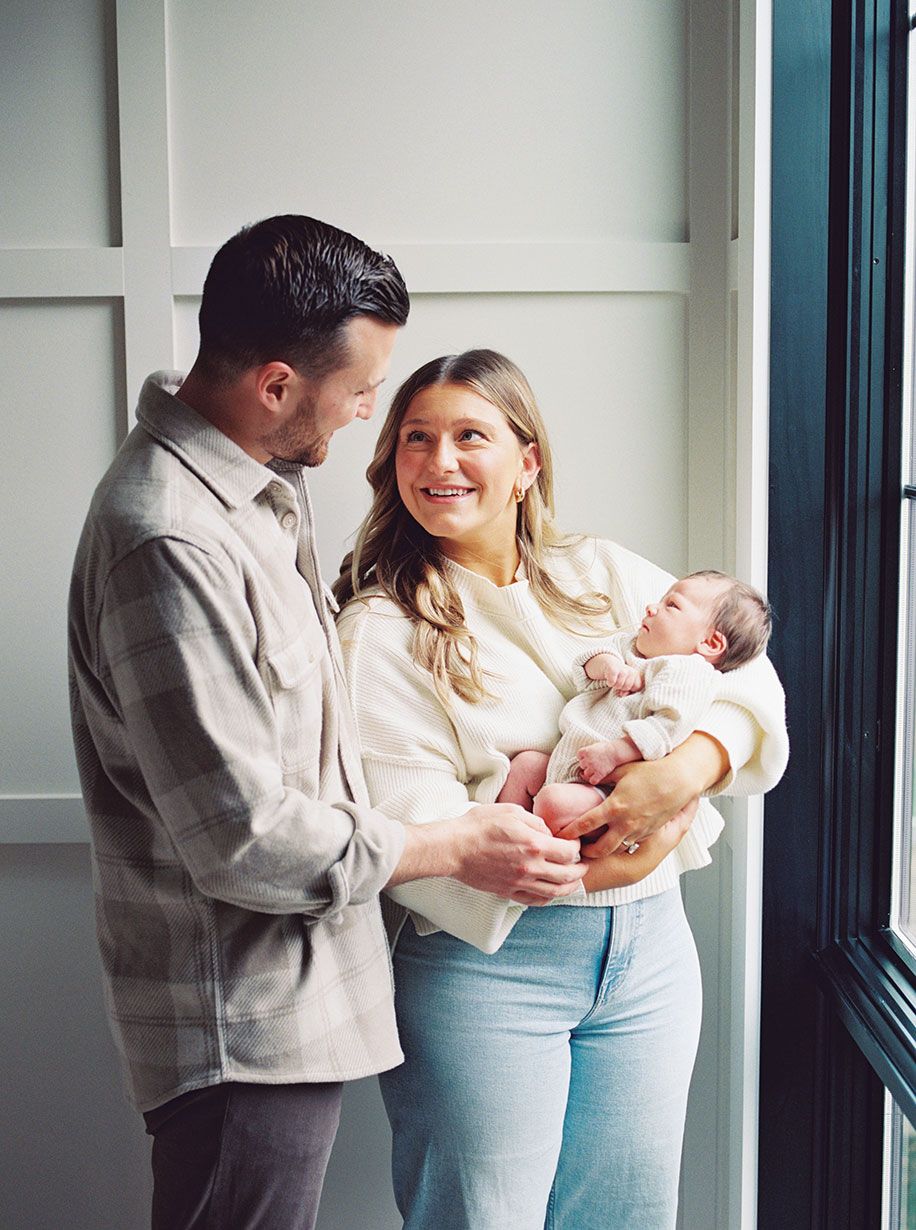 A couple stands by a window sharing a tender moment with their newborn baby in a bright, modern interior.