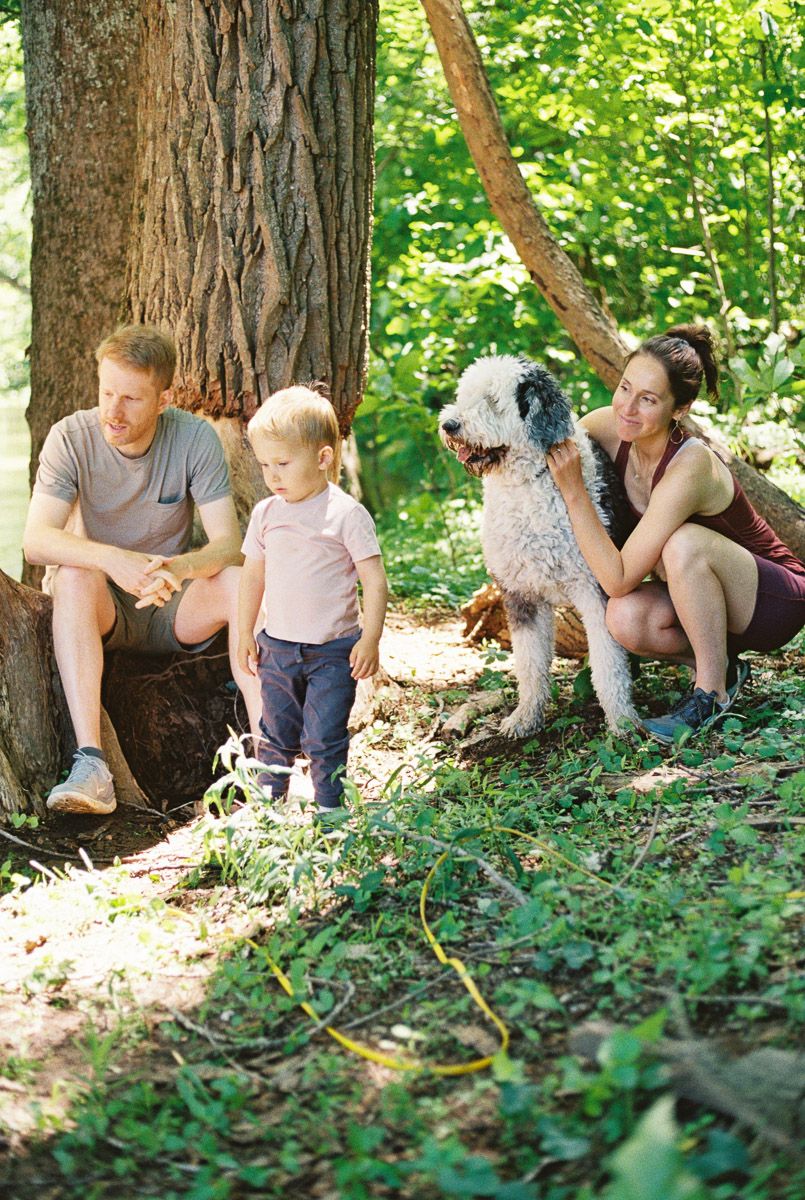 Family with toddler and fluffy dog relaxing in a sunny forest near a large tree trunk.
