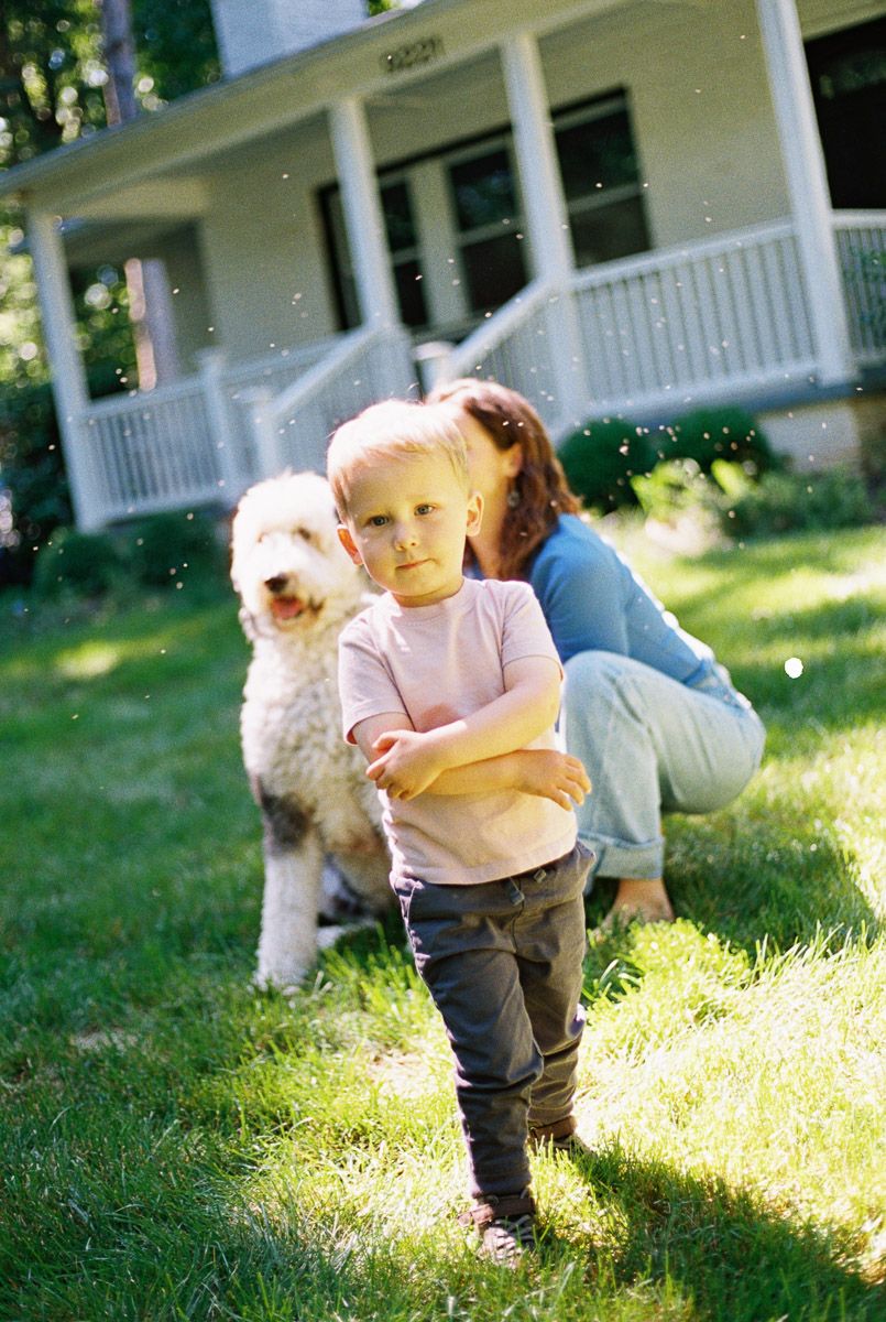 Young child with crossed arms stands on grass beside a fluffy white dog and a woman near a white house.