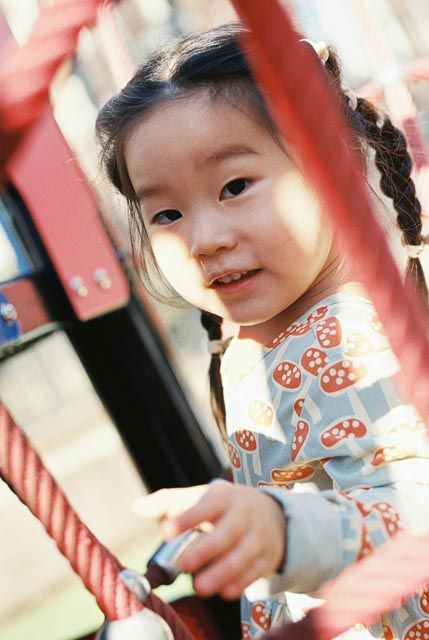 Smiling Asian toddler girl with braided pigtails playing on red playground structure.