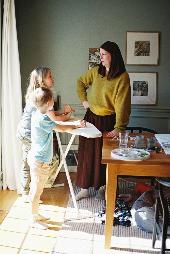 Child in light blue shirt and yellow pants reaches for papers on dining table while adult in yellow sweater assists.