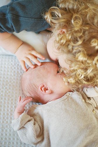 Curly blonde toddler gently kissing a newborn baby sibling lying on a soft white blanket.