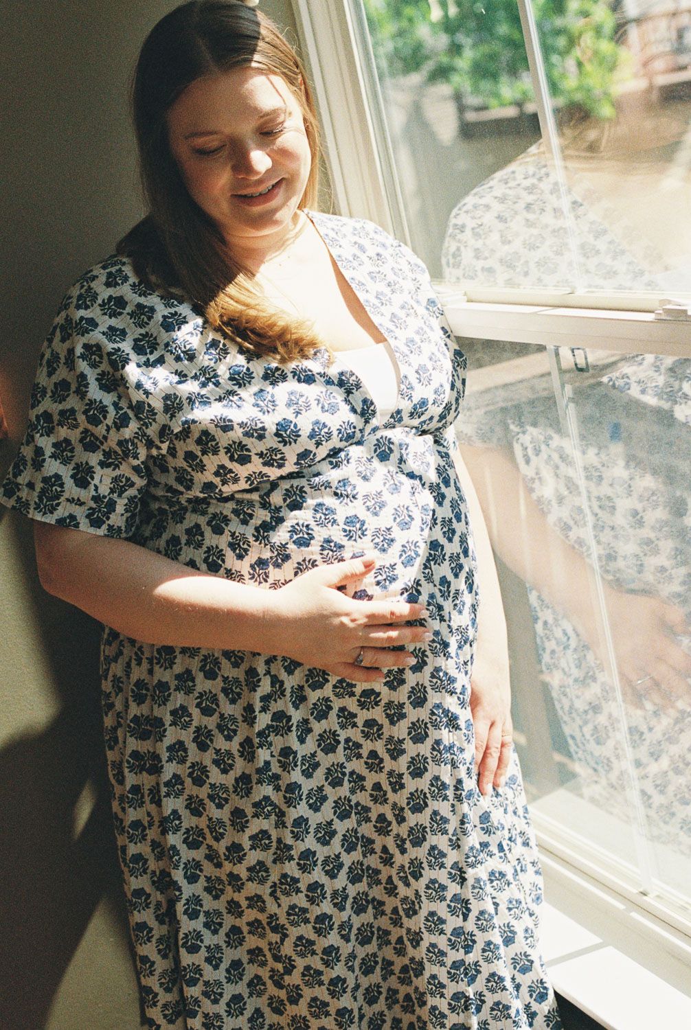 Pregnant woman in floral blue and white wrap dress cradling baby bump by sunlit window, smiling softly.