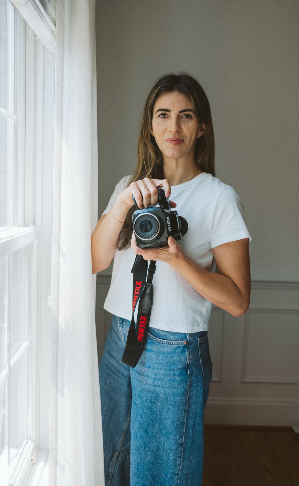 Photographer in white t-shirt and jeans holds professional camera by bright window in minimalist indoor setting.