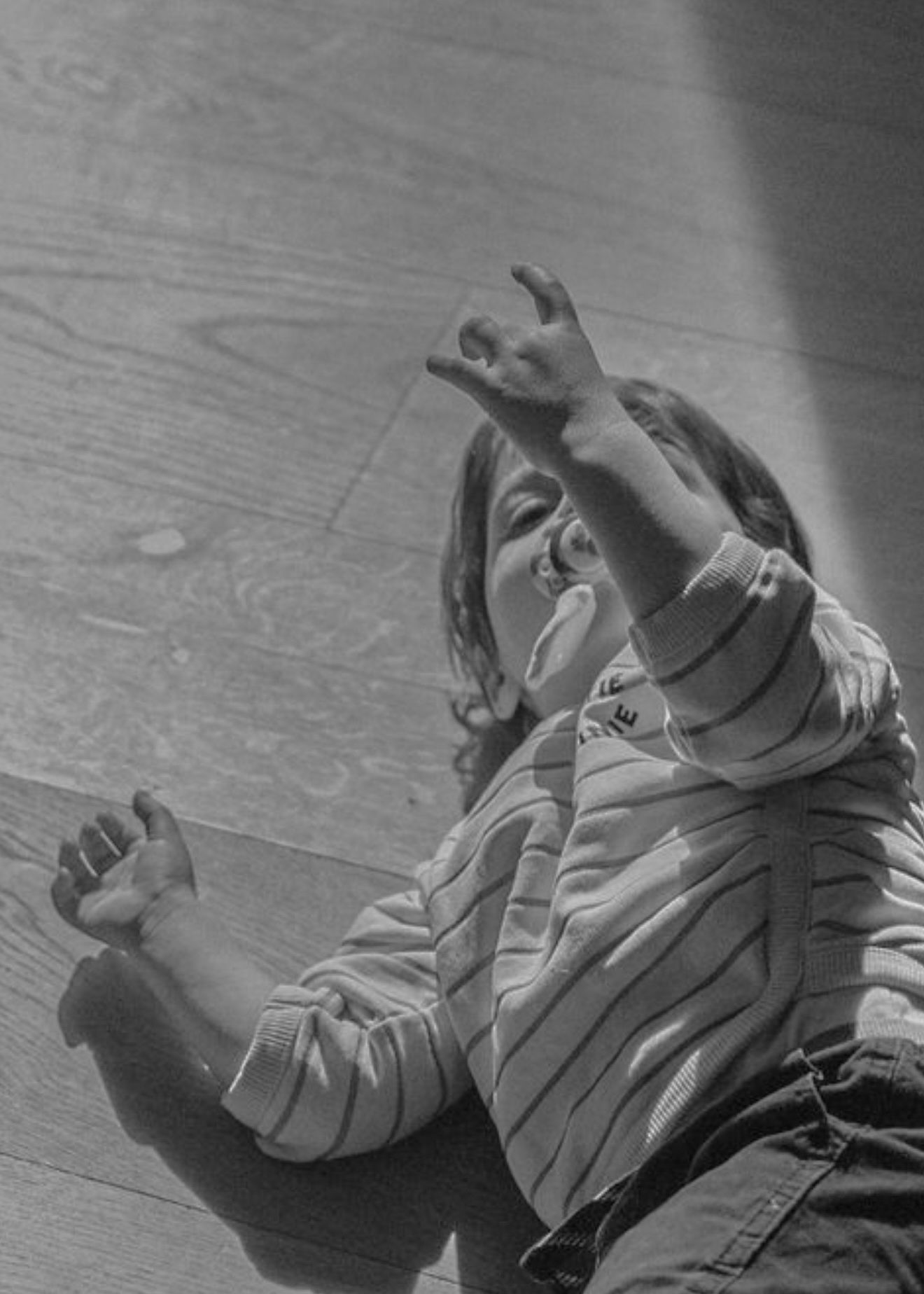 A small child lying on a hardwood floor reaches up joyfully with one arm in this black and white photograph.