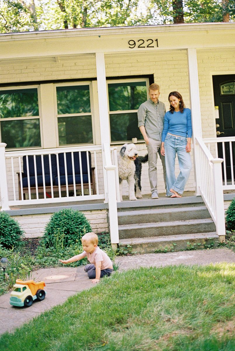 Family with dog stands on porch of house 9221 while toddler plays with toy truck on the sidewalk.