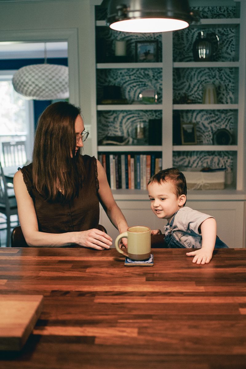 A mother and toddler share a warm moment at a wooden dining table with a yellow mug in a cozy home kitchen.